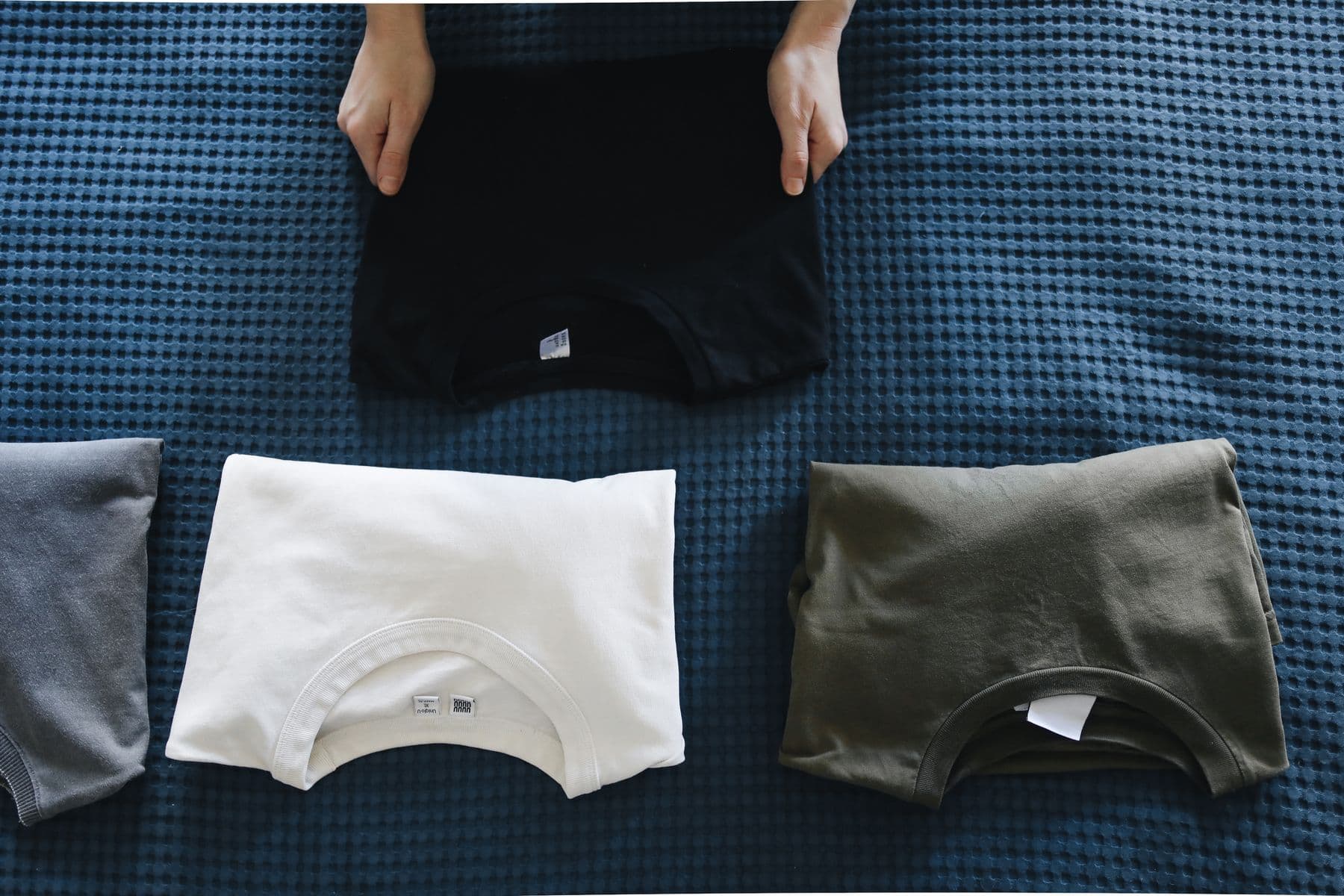 Hände falten T-Shirts zu ordentlichen Stapeln auf einer Decke (Symbolbild)