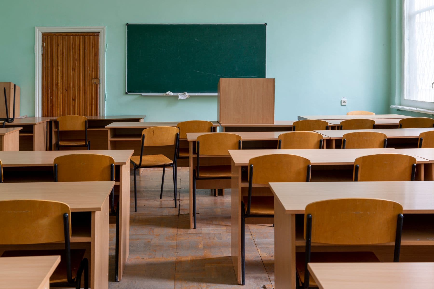 Ein leeres Klassenzimmer mit Holztischen, Stühlen und einer grünen Tafel.